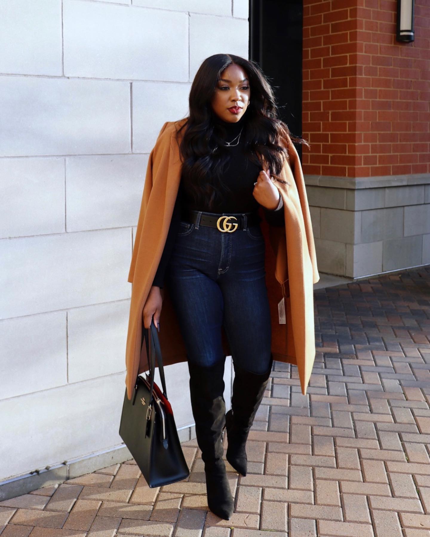 Jaslyn outdoors in a camel coat, dark jeans, and tall black boots, carrying a dark handbag